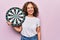 Middle age beautiful woman holding dartboard standing over isolated pink background looking positive and happy standing and