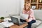 Middle age blonde woman teleworking using laptop and talking on the smartphone at home