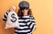 Middle age brunette burglar woman wearing mask and cap holding bag with dollar symbol covering mouth with hand, shocked and afraid
