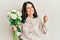 Middle age brunette woman holding bouquet of white flowers screaming proud, celebrating victory and success very excited with