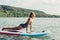 Middle age Caucasian woman practising yoga on paddle sup surfboard at sunset. Female stretching doing workout on a lake water.