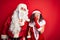 Middle age couple wearing Santa costume and glasses over isolated red background hand on mouth telling secret rumor, whispering