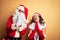 Middle age couple wearing Santa costume and glasses over isolated yellow background begging and praying with hands together with