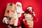 Middle age couple wearing Santa costume holding tower of gifts over isolated red background hand on mouth telling secret rumor,