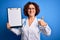 Middle age curly hair doctor woman wearing coat and stethoscope holding clipboard happy with big smile doing ok sign, thumb up