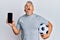 Middle age grey-haired man holding football ball showing smartphone angry and mad screaming frustrated and furious, shouting with