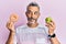 Middle age grey-haired man holding green apple and donut smiling looking to the side and staring away thinking