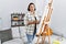 Middle age hispanic woman drawing by painter easel stand at home thinking attitude and sober expression looking self confident