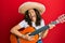 Middle age hispanic woman holding mexican hat playing classical guitar smiling and laughing hard out loud because funny crazy joke