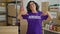 Middle age hispanic woman pointing to volunteer uniform smiling at charity center