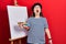 Middle age hispanic woman standing drawing with palette by painter easel stand angry and mad screaming frustrated and furious,