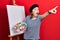 Middle age hispanic woman standing drawing with palette by painter easel stand pointing with finger surprised ahead, open mouth