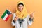 Middle age hispanic woman wearing hijab holding kuwait flag smiling happy pointing with hand and finger to the side