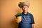 Middle age hoary farmer man wearing apron and hat holding plant pot over yellow background scared in shock with a surprise face,