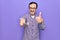 Middle age man changing tv channel using television remote control over purple background smiling happy and positive, thumb up