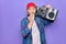 Middle age man wearing cap listening vintage boombox over isolated purple background covering mouth with hand, shocked and afraid