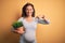 Middle age pregnant woman expecting baby holding plant pot with surprise face pointing finger to himself