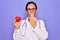 Middle age senior cardiologist doctor woman holding red heart over purple background cover mouth with hand shocked with shame for