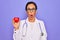 Middle age senior cardiologist doctor woman holding red heart over purple background scared in shock with a surprise face, afraid