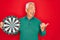 Middle age senior grey-haired man holding competition dartboard target over red background pointing and showing with thumb up to