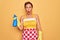Middle age senior housewife pin up woman wearing 50s style retro dress cleaning using spray scared in shock with a surprise face,