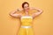 Middle age senior pin up woman wearing 50s style retro dress over yellow background relaxing and stretching, arms and hands behind