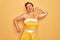 Middle age senior pin up woman wearing 50s style retro dress over yellow background stretching back, tired and relaxed, sleepy and