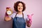 Middle age woman cleaning doing housework wearing apron and gloves holding alarm clock pointing and showing with thumb up to the