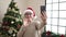 Middle age woman with grey hair having video call standing by christmas tree at home