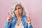 Middle age woman with grey hair standing over pink background pointing up looking sad and upset, indicating direction with