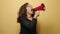 Middle age woman screaming angry using megaphone over yellow background