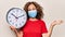 Middle age woman wearing coronavirus protection mask holding big clock ticking for deadline celebrating achievement with happy