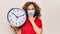 Middle age woman wearing coronavirus protection mask holding big clock ticking for deadline covering mouth with hand, shocked and