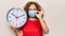 Middle age woman wearing coronavirus protection mask holding big clock ticking for deadline doing ok sign with fingers, smiling