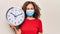 Middle age woman wearing coronavirus protection mask holding big clock ticking for deadline looking positive and happy standing