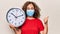 Middle age woman wearing coronavirus protection mask holding big clock ticking for deadline pointing thumb up to the side smiling