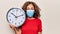 Middle age woman wearing coronavirus protection mask holding big clock ticking for deadline scared and amazed with open mouth for