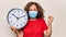 Middle age woman wearing coronavirus protection mask holding big clock ticking for deadline screaming proud, celebrating victory