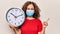 Middle age woman wearing coronavirus protection mask holding big clock ticking for deadline smiling happy pointing with hand and