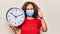 Middle age woman wearing coronavirus protection mask holding big clock ticking for deadline smiling happy and positive, thumb up