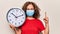 Middle age woman wearing coronavirus protection mask holding big clock ticking for deadline smiling with an idea or question