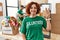 Middle age woman wearing volunteer t shirt at donations stand showing and pointing up with fingers number three while smiling