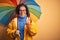 Middle age woman wearing yellow raincoat under colorful umbrella over isolated background happy with big smile doing ok sign,