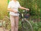 Middle-aged attractive slim woman in light trousers and shirt stands near the bike in the Park on a Sunny summer day, Cycling
