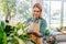 Middle-Aged Businesswoman in Apron, Tenderly Dusting Plant Leaves in Flower Shop, Seamlessly Directing a Floristry