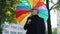 Middle-aged caucasian grey-haired man wearing glasses and blazer standing at park with rainbow umbrella looking around