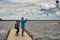 Middle-aged couple standing on the wooden jetty, photographing and filming kitesurfers on the lake