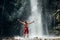 Middle-aged man dressed only red trekking shorts standing under the mountain river waterfall, rose arms up and enjoying the