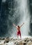 Middle-aged man dressed only red trekking shorts standing under the mountain river waterfall, rose arms up and enjoying the