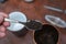 Middle-aged man holding spoonful of dry tea. Crushed black tea leaves in spoon over red table. Cup and container are blurred on